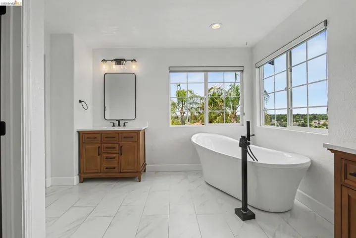 Bathroom featuring a freestanding soaking tub with a matte black floor-mounted filler, large grid-pattern windows, a wood-finish vanity with a white countertop and rectangular sink, a black framed mirror, and marble-look floor tiles