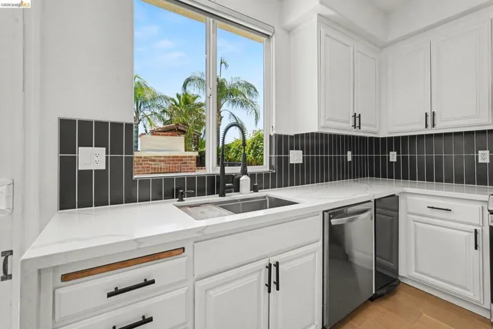Kitchen featuring white shaker-style cabinetry, a stainless steel double basin sink, black vertical tile backsplash, light-toned countertops, and a stainless steel dishwasher