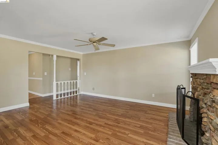 Spacious room featuring wood-finish flooring, a stone-front fireplace with a brick hearth, crown molding, and a ceiling fan