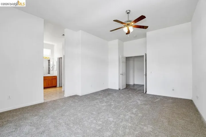 Spacious room featuring neutral carpeting, a ceiling fan with integrated lighting, and direct access to a separate area with tiled flooring and a wood vanity