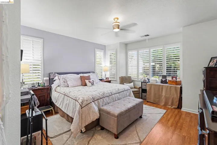 Spacious bedroom featuring wood-finish flooring, a ceiling fan, and multiple windows with plantation shutters