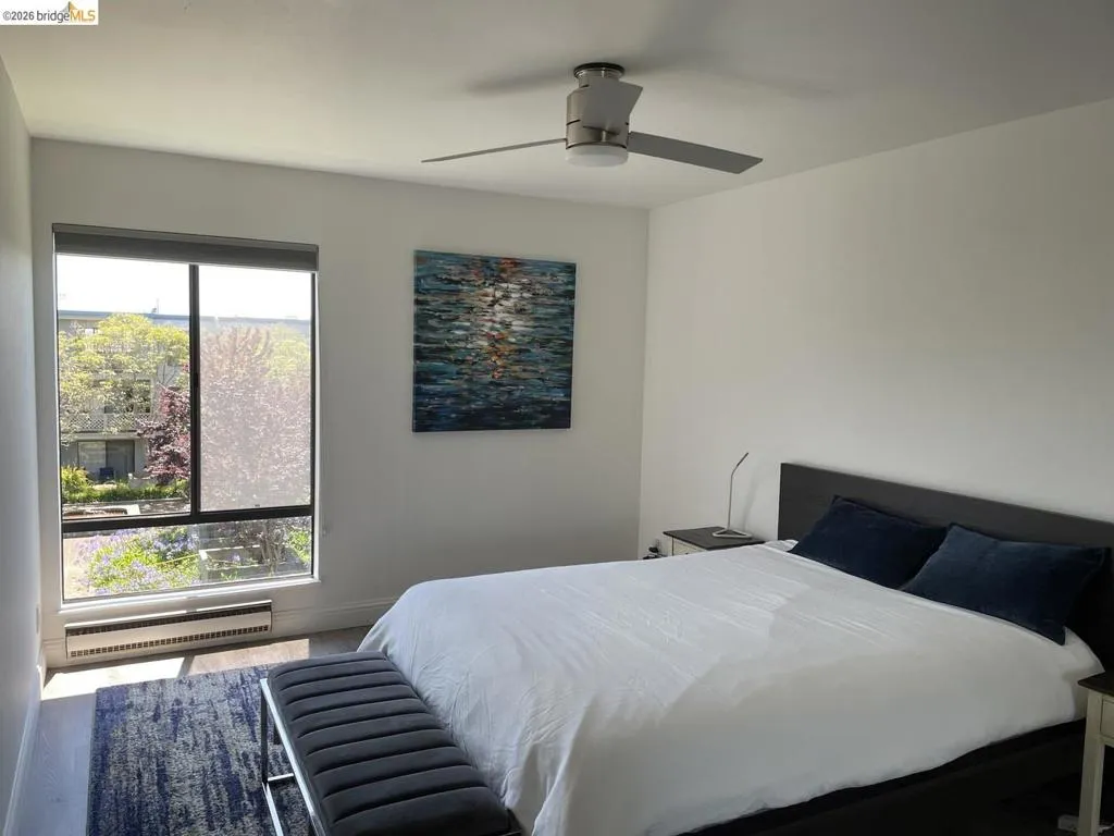 Bedroom featuring a large multi-pane window with roller shade, wood-finish flooring, and a ceiling fan with integrated lighting