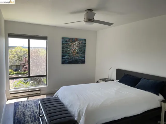 Bedroom featuring a large multi-pane window with roller shade, wood-finish flooring, and a ceiling fan with integrated lighting