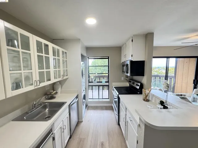 Galley kitchen featuring light wood-finish flooring, white cabinetry with glass-front uppers, stainless steel appliances, and solid surface countertops