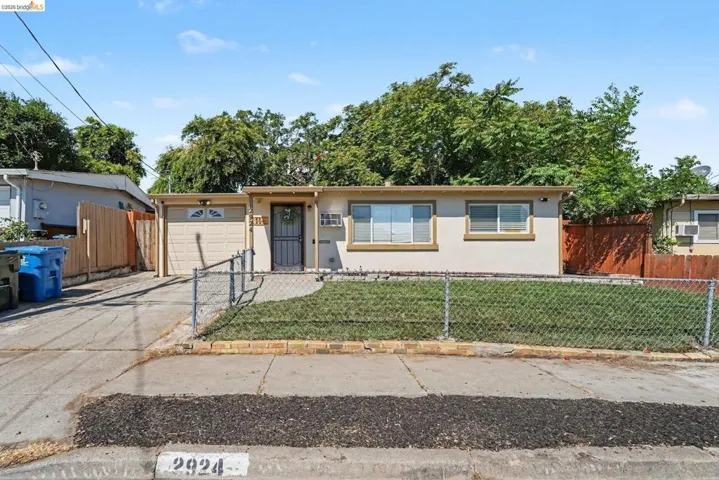Single-story stucco residence featuring a chain-link fence, a front lawn, and a paved driveway leading to an attached garage