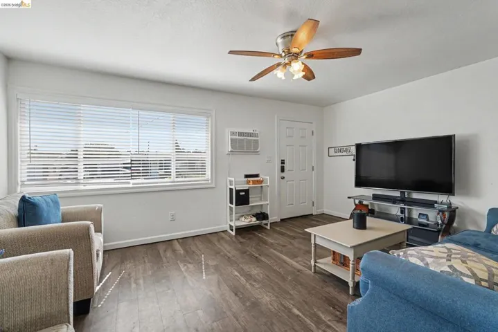 Living area featuring wood-finish flooring, a multi-pane window with horizontal blinds, a white paneled entry door, and a ceiling fan with light fixture