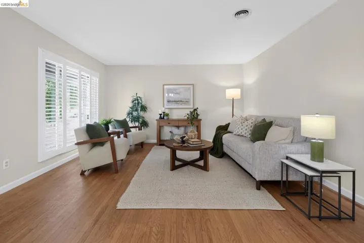 Spacious room with wood-finish flooring, light-toned wall paint, white louvered window treatments, and a ceiling-mounted ventilation register