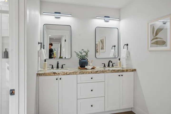 Double vanity featuring a light-toned stone countertop, white cabinetry with black hardware, twin rectangular mirrors with black frames, and modern linear lighting fixtures