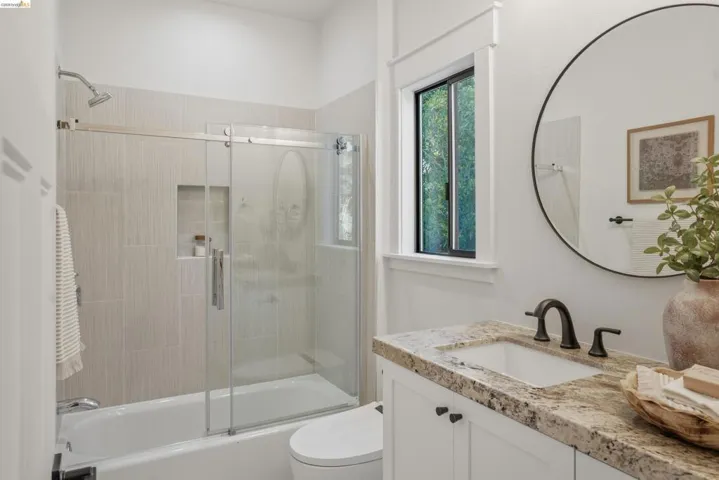 Bright bathroom featuring a bathtub with a sliding glass enclosure, light-toned wall tiling, a built-in shower niche, a window with dark trim, and a vanity with a stone countertop