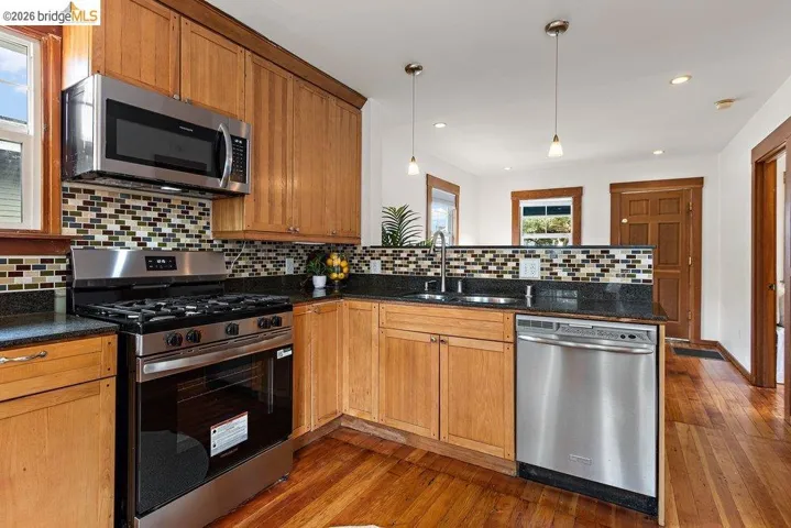 Kitchen featuring light wood cabinetry, stainless steel appliances, dark countertops, a mosaic tile backsplash, and wood-finish flooring