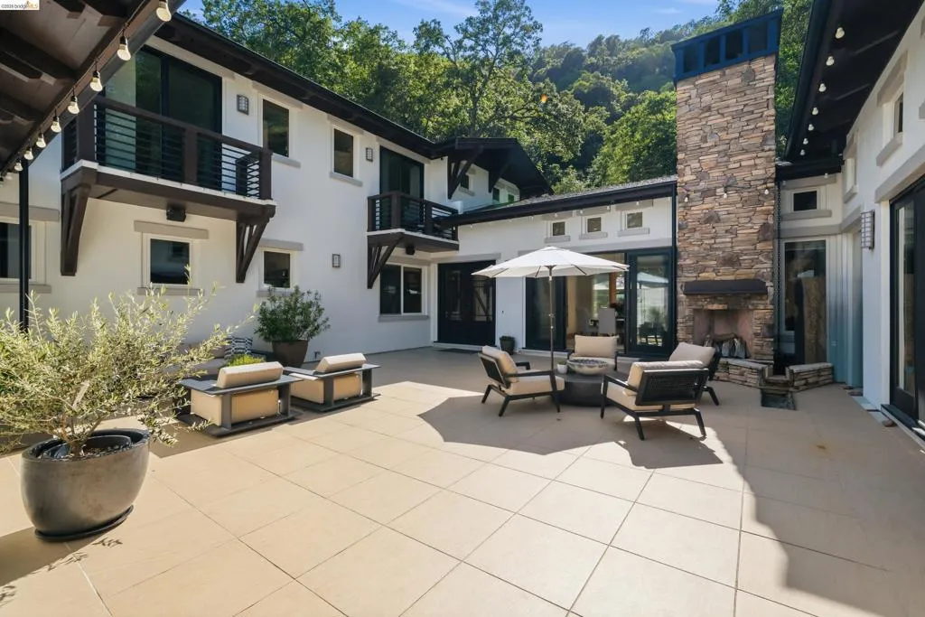 Expansive outdoor patio featuring large format tile flooring, a floor-to-ceiling stacked stone fireplace, multiple balconies with dark wood railings, and black framed glass doors