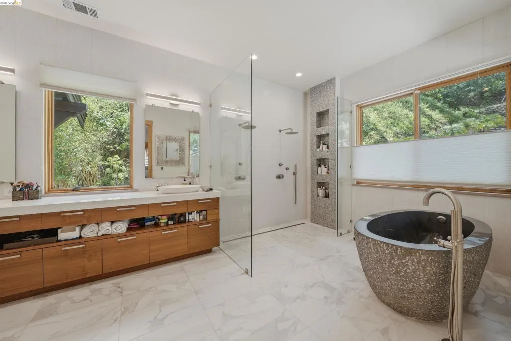 Spacious bathroom featuring a freestanding stone soaking tub, a large glass-enclosed shower with multiple showerheads, a dual vanity with wood-finish cabinetry, and wide windows