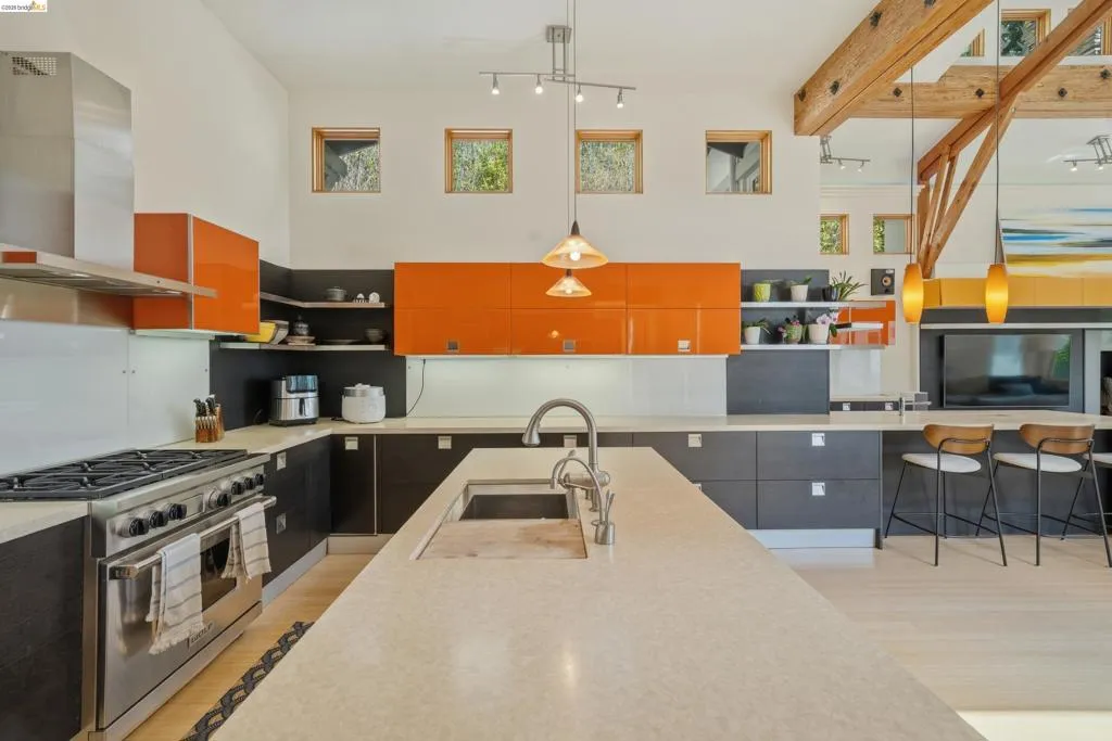 Expansive kitchen featuring a large central island with an undermount sink, stainless steel range with a professional hood, two-tone cabinetry with orange accents, and natural wood ceiling beams