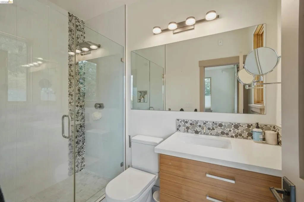 Modern bathroom featuring a frameless glass shower enclosure, a full vanity mirror, a wood-finish vanity with an integrated sink, a mosaic tile backsplash, and a contemporary light fixture