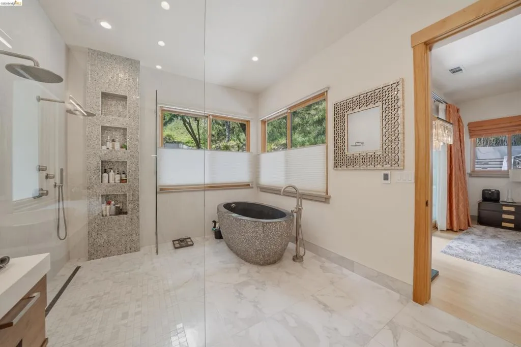 Spacious bathroom featuring a freestanding stone soaking tub, a walk-in shower with dual showerheads and recessed shelving, wood-framed windows, and recessed lighting