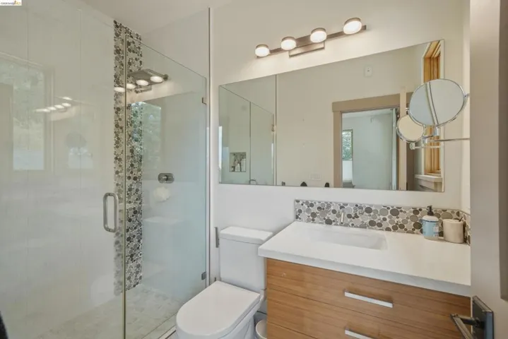 Modern bathroom featuring a frameless glass shower enclosure, a full vanity mirror, a wood-finish vanity with an integrated sink, a mosaic tile backsplash, and a contemporary light fixture