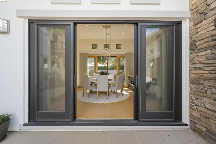 Sliding glass doors with dark frames open to an interior dining area featuring wood-finish flooring, a contemporary chandelier, and recessed lighting