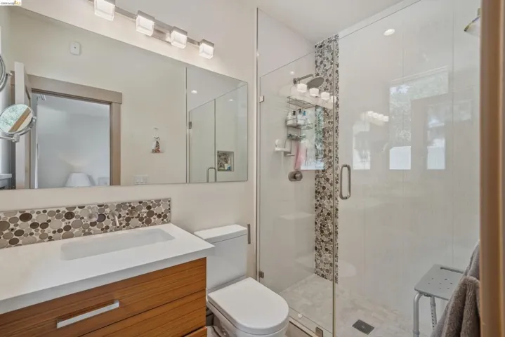 Full bathroom featuring a glass-enclosed shower with a mosaic tile accent strip, a floating wood-finish vanity with an integrated rectangular sink, a white countertop, and a full-width mirror with linear vanity lighting