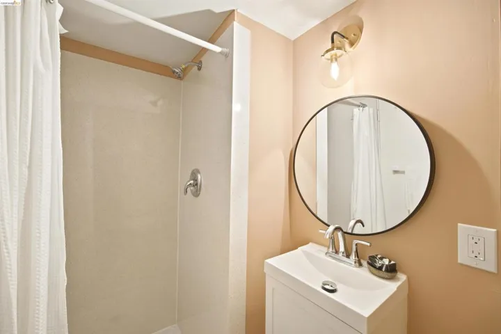 Compact bathroom featuring a shower with light-toned paneling, a white vanity with an integrated sink, a circular mirror with a dark frame, a brass-finished wall sconce, and polished chrome fixtures