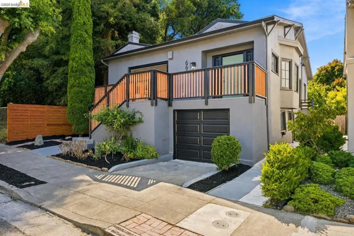 Contemporary stucco exterior featuring a built-in garage with a paneled door, a second-story balcony with wood and metal railing, established landscaping, and a wood privacy fence