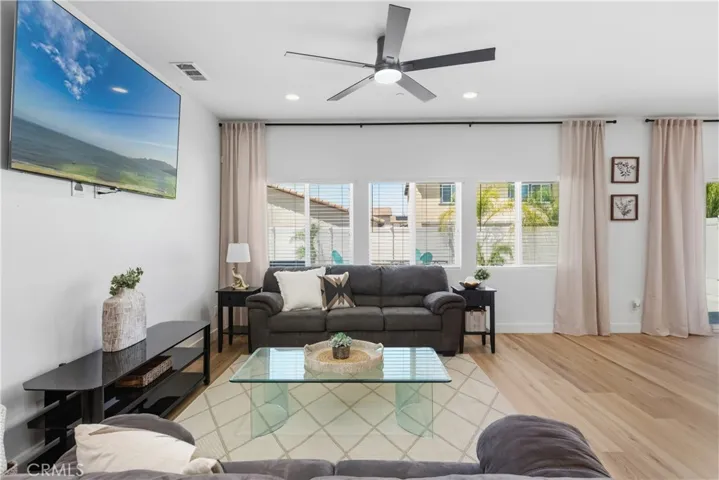 Spacious great room showcasing wide-plank natural tone laminate flooring, black modern ceiling fan, abundant natural light from picture windows and glass slider, plus black curtain rods with linen drapes