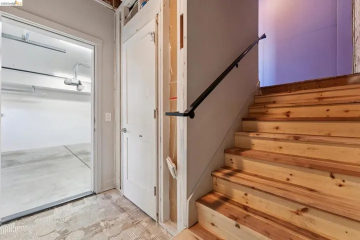 Exposed wood staircase with a black metal handrail, featuring an adjacent white paneled door and an open doorway to a garage space with concrete flooring and overhead lighting