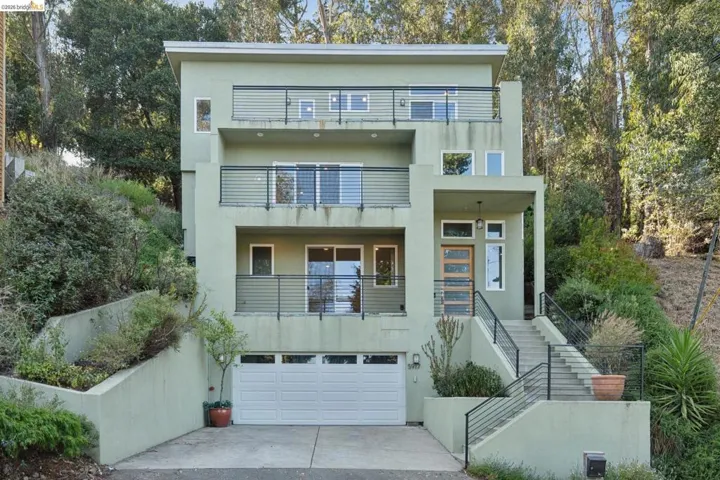Modern multi-level residence featuring a sage green exterior, multiple balconies with horizontal railings, and a prominent front entrance with a contemporary wood-paneled door