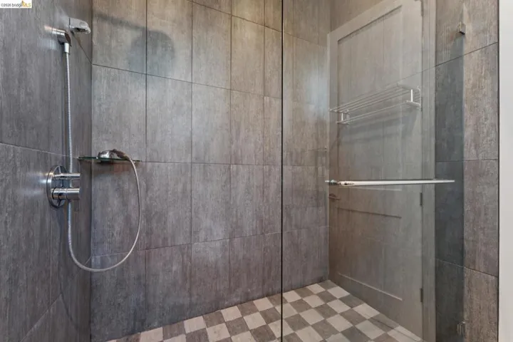 Walk-in shower featuring full-height gray tile walls, a checkerboard tile floor, a chrome shower system with a handheld sprayer, and a frameless glass enclosure with a bar handle