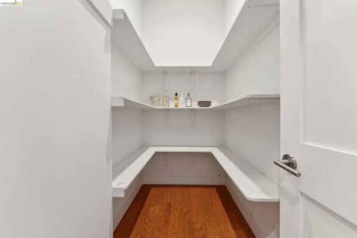 Walk-in pantry featuring white shelving, wood-finish flooring, and a contemporary door handle