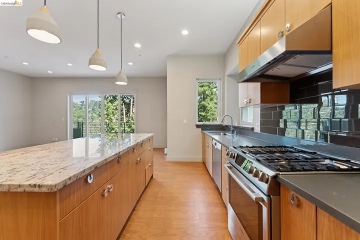 Modern kitchen featuring a large granite-topped island with pendant lighting, wood-finish cabinetry, stainless steel appliances, and a dark tile backsplash