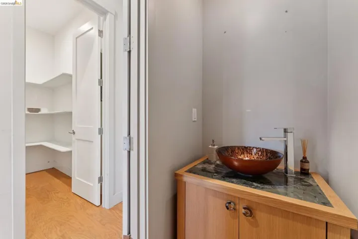 Powder room featuring a vessel sink with a contemporary faucet, wood-finish vanity with stone countertop, and an adjacent pantry with wood-finish flooring and built-in shelving