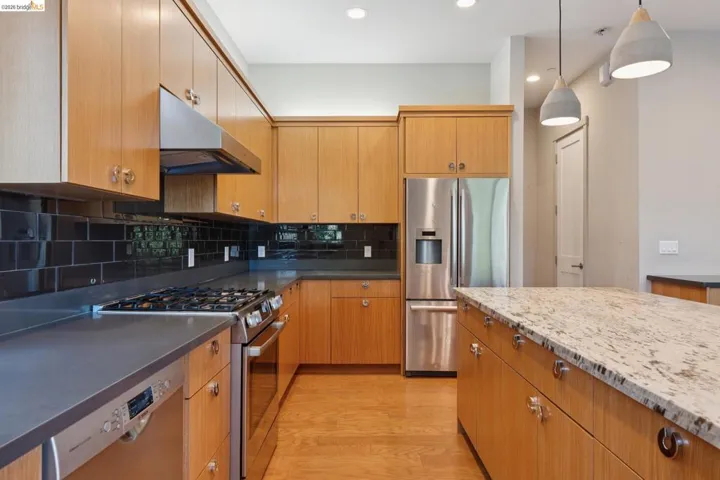 Kitchen featuring wood-finish cabinetry, stainless steel appliances, dark subway tile backsplash, and a central island with a light-toned countertop