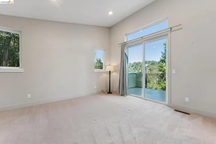Spacious room featuring light-toned carpeting, high ceilings, multiple windows, and a large sliding glass door opening to an exterior balcony