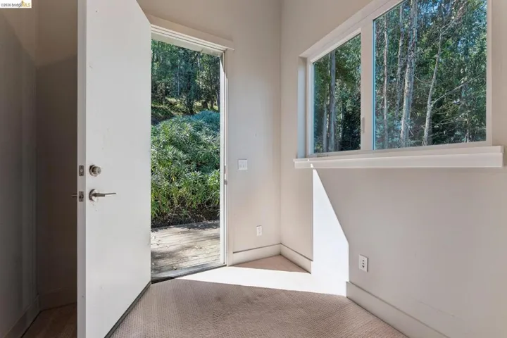 Interior room featuring a white door opening to a wooded exterior, a large window with white trim, and light beige carpet flooring