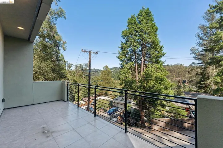 Outdoor balcony featuring light-colored tile flooring, sleek black horizontal railings, recessed overhead lighting, and abundant surrounding tree canopy