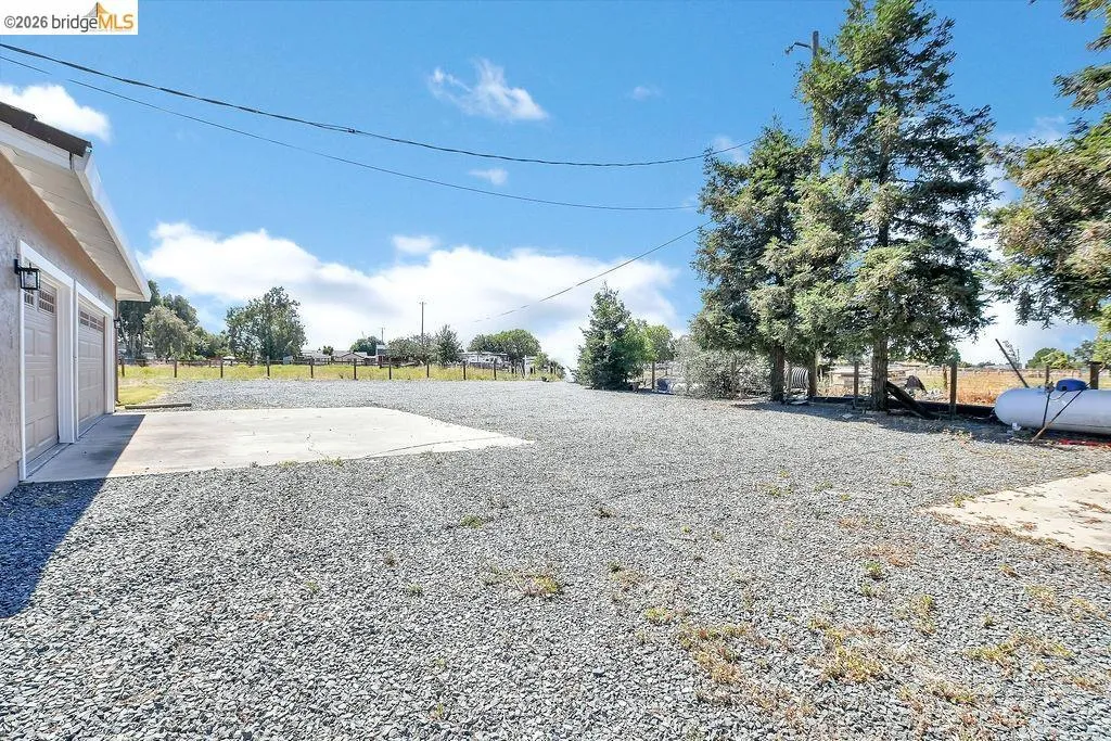 Expansive gravel lot featuring a two-car garage with overhead doors, a concrete pad, and mature evergreen trees