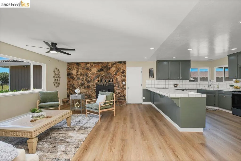 Open-concept living area featuring wood-finish flooring throughout, a stone accent wall with a fireplace, and a contemporary ceiling fan