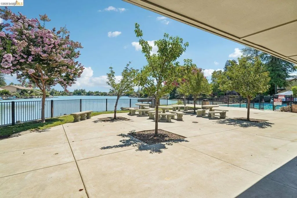 Waterside patio area featuring multiple picnic tables with benches, a black metal fence bordering the water, and mature trees providing shade