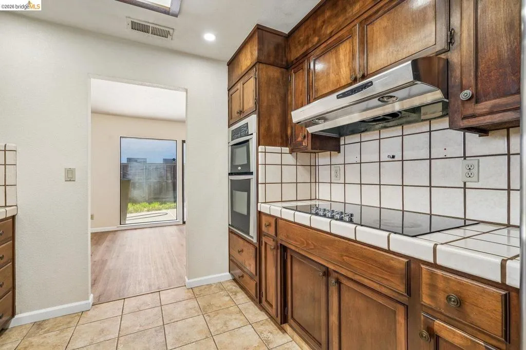 Kitchen featuring wood cabinetry, white tile countertops, a built-in electric cooktop, a stainless steel range hood, and double wall ovens