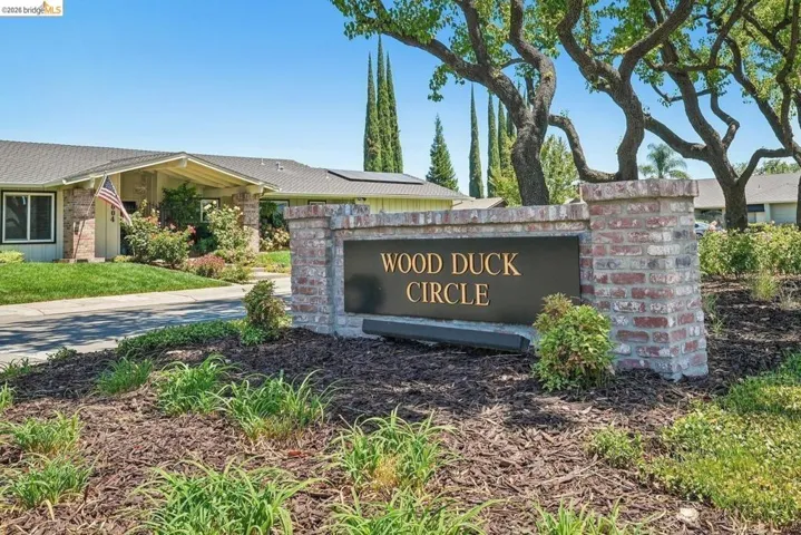 Brick-clad monument sign with "WOOD DUCK CIRCLE" lettering, set within a landscaped area featuring mature trees and ground cover