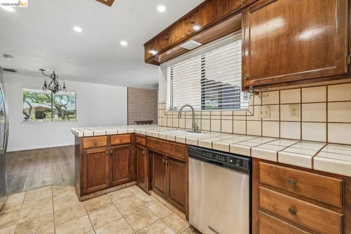 Kitchen featuring wood cabinetry, tile countertops, stainless steel dishwasher, and a window with blinds