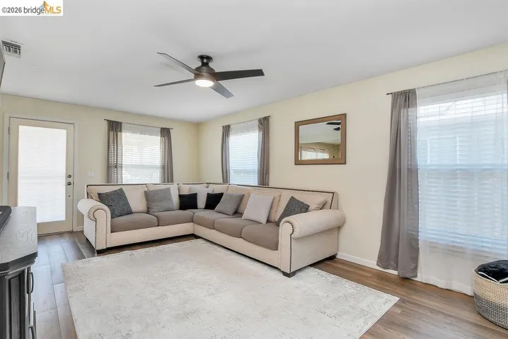 Living area featuring vinyl flooring, light-toned walls, multiple windows with blinds and drapes, and a ceiling fan with integrated lighting