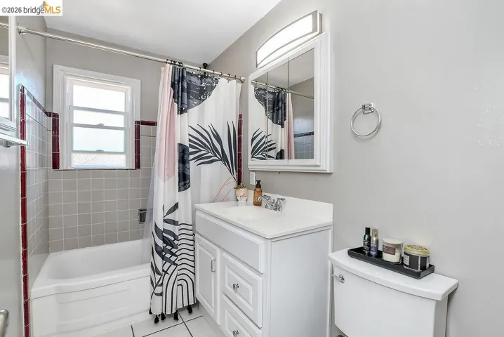 Full bathroom featuring a white vanity with integrated sink, white tiled flooring, a built-in bathtub with light gray square tiles and a dark red trim, and a window with frosted glass