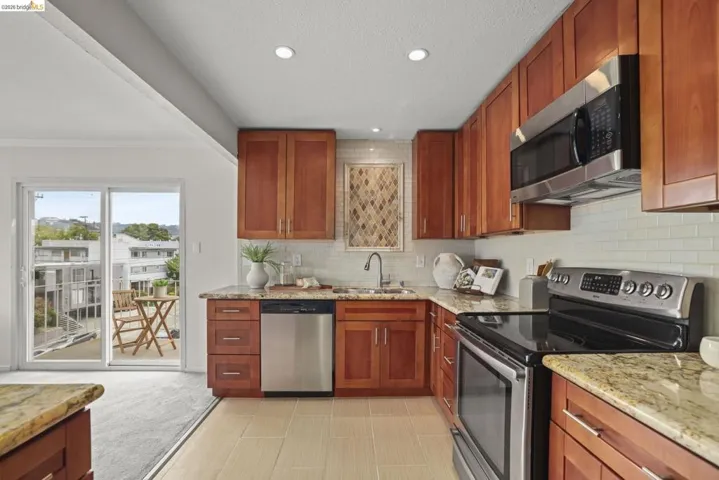Modern kitchen featuring wood cabinetry, stainless steel appliances, light-toned tile flooring, and light granite countertops
