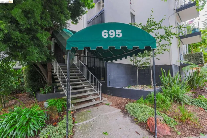 Building entrance featuring a green awning with address numbers, a concrete pathway, a multi-level staircase with white handrails, and surrounding mature landscaping