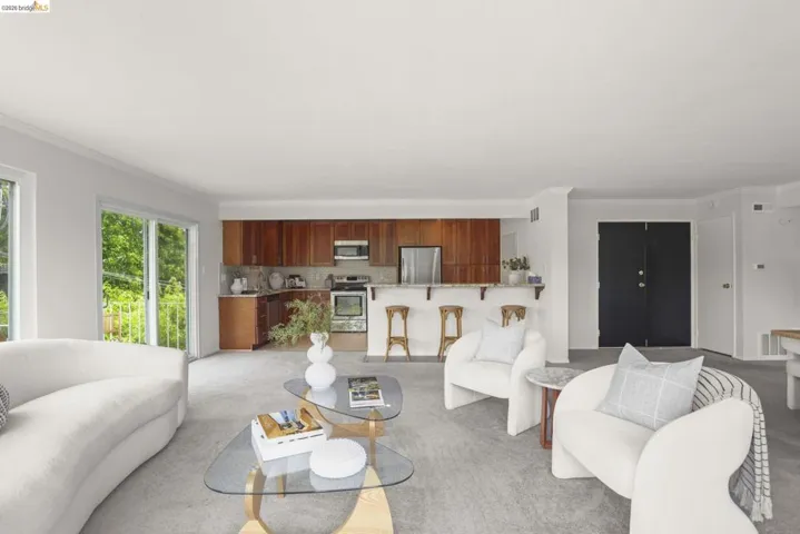 Open-concept living space featuring light gray carpeting, white walls, and a kitchen area with wood cabinetry, stainless steel appliances, and a breakfast bar with a light-toned countertop