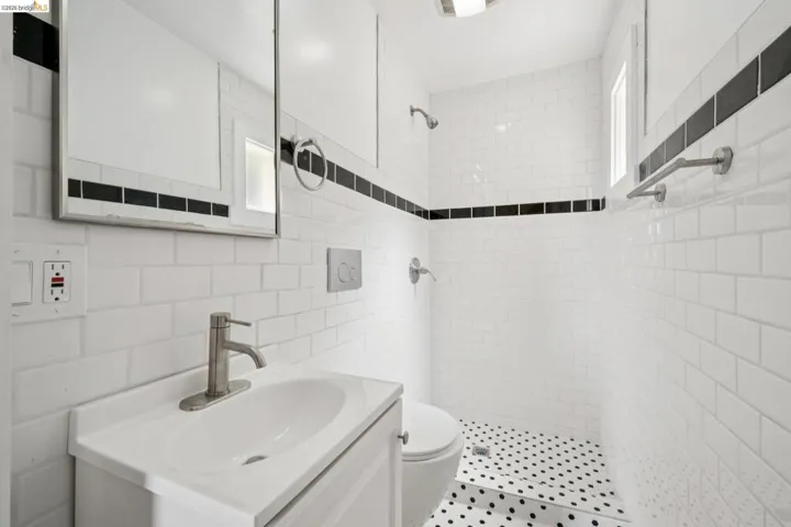 Bathroom featuring subway tile walls with black accent banding, a white vanity with an integrated sink, a mirrored medicine cabinet, a wall-mounted toilet, and a shower with black and white penny tile flooring