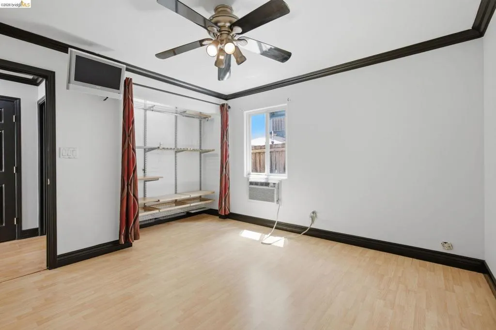 Room featuring wood-finish flooring, dark trim, a ceiling fan with integrated lighting, a window-mounted air conditioning unit, and built-in shelving with hanging space