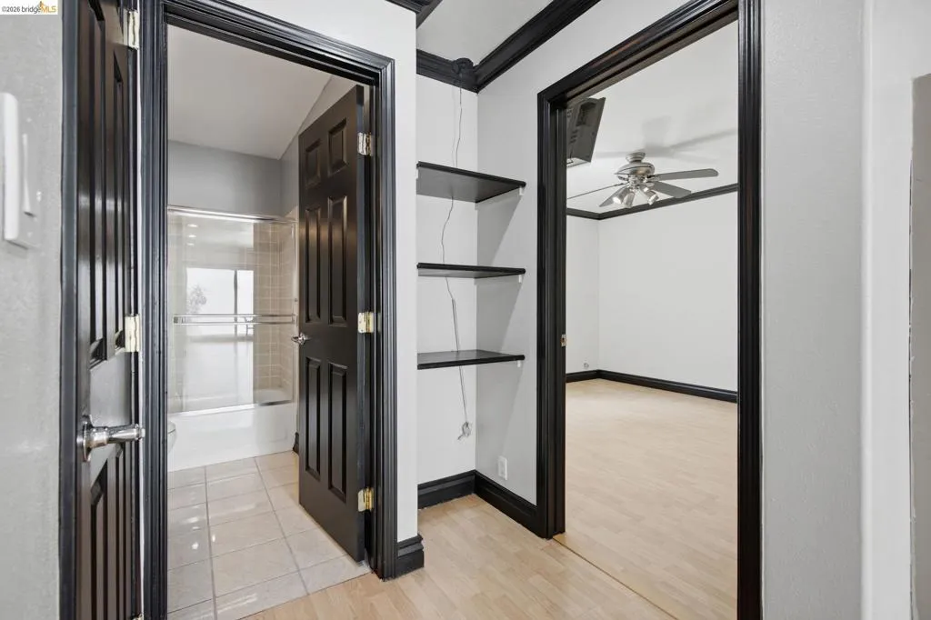 Hallway featuring light wood-finish flooring, dark trim and doors, built-in shelving, and entryways to a room with a ceiling fan and a bathroom with a shower-over-tub combination and tiled flooring