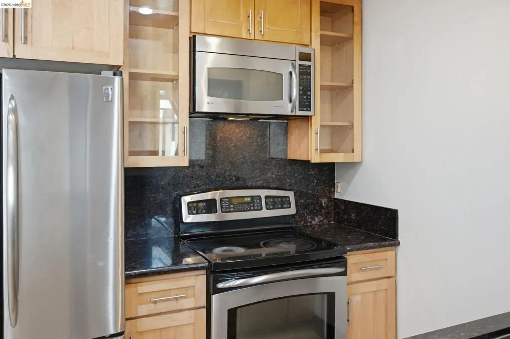 Kitchen featuring light wood-finish cabinetry, dark stone countertops, stainless steel appliances, and a matching stone backsplash